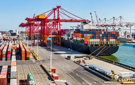 Photo of a port with a large carrier container ship filled up and containers on the pavement near it.